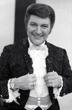 Grayscale medium shot of Liberace smiling in sequined clothes making eye contact with the camera lens. His hands, adorned with two rings and a bracelet, hold the lapel of his sequined suit. Like the suit, the bowtie and vest appear to sparkle.