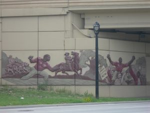 An elongated mural on the I-43 overpass wall in Milwaukee displays caricatural images of people running from slavery. Drawn on the wall, is a figure of a man holding a protest sign that reads "Free Joshua Glover Now" while parading Glover on his shoulder.