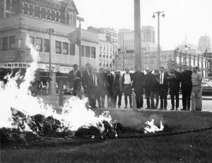 Long shot of fourteen men standing arm-in-arm on the center-to-right in grayscale. A blazing fire from burned draft cards is on lower ground in the image's foreground. Milwaukee's buildings soar in the far background.
