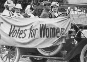 Grayscale photograph of members of the Political Equality League sitting in an early model Ford car running to the right. The women appear in hats. A man in a beret hat is seated in the driver's seat. The automobile's side is draped with a banner that reads "Votes for Women." The exterior wall of a building is visible in the background.