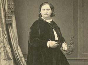 A full body shot of Mathilde Franziska Anneke in a studio wearing a long, dark velvet skirt and jacket covering her legs and arms. She poses in a standing position with a pen in her right hand and a small book her her left. Her face looks to her right.