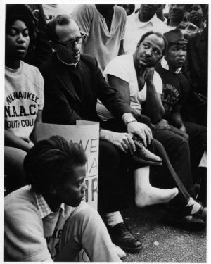 Grayscale full shot of Father James Groppi sitting with a group of young people in his glasses and clergy shirt. He sits in the middle while putting on his left shoe. Some of the marchers wear sweatshirts that read "Milwaukee N.A.A.C.P Youth Council."