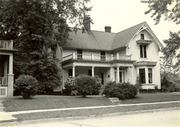 Grayscale wide shot of the Beulah Brinton House facade in a residential area. The L-framed building consists of two-and-a-half-stories. The narrow side of the structure is on the right featuring windows on each of its stories. The longer side on the left has an entrance on the ground floor and a balcony on the second floor enclosed by balustrades.