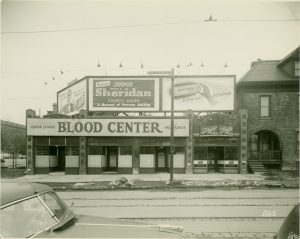 Sepia-colored image of the Junior League's Blood Center facade by Wells Street. The place features three entrance doors, as it consists of three shops combined. Attached along the building's top is the rectangular "Junior League Blood Center" sign. The storefront is adjacent to a Schlitz beer tied house. Above their flat roof are billboards advertising Schlitz beer, a county judge's reelection campaign, and a chewing gum brand.