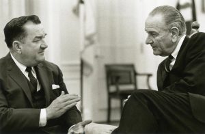 Grayscale medium shot of Representative Zablocki on the left while speaking with President Lyndon B. Johnson on the right. They appear in suits and ties. Both face each other. A blurry image of a flag and a chair are visible in the background.