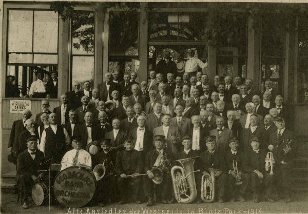 Group photo of George Bach Jr.'s Band and Orchestra in sepia. Dozens of the members pose on the staircase and the front of a building. Only the first row is seated while posing with a drum and various brass instruments. The group's name is inscribed on the bass drum shield. Some people at the back stand at the building's entrance. Some smile from inside a window on the left foreground.