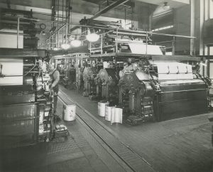 Grayscale photograph of the interior of Milwaukee Journal press room. A man works on a machine on the left. He stands on an aisle. Several brightly lit lamps are hung in a row above the aisle. Behind the man is a row of equipment. Steel structures and concrete walls are visible.