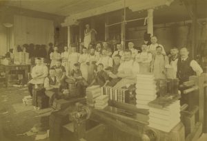 Sepia-colored group photo of dozens of Brumder Publishing Company employees posing while making eye contact with the camera lens. Some younger workers in the front sit on top of stacks of books. Others stand behind in several rows while crossing their arms. The ceiling and posts are visible in the background. A man hugs a post while standing higher than anybody in the room. Book stacks appear in the right foreground and left background.
