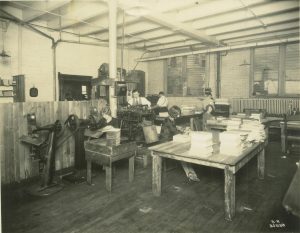 Sepia-colored image of people binding books for the Krueger Printing Company. Two machines sit on the left next to a wooden partition that divides part of the room. A woman works on one of them. Behind her is a man working with another book-binding machine. Beside the woman are other female employees sitting at two tables full of book stacks. Their bodies face to the right. The backs of two men working on machines appear in the background, along with the brick walls, windows, and ceiling lights.