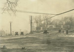 A sepia-colored long shot of the unpaved intersection of Hawley Road and State Street. Automobiles in the background traverse the street. Two cars appear on the left, and one turns right onto an uphill road. Utility poles and billboards are visible on the street corners.