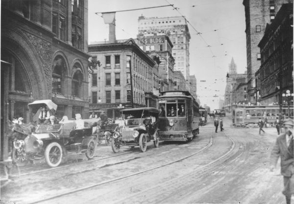 Grayscale long shot of automobiles, horse-drawn carriages, and trolleys traversing E. Wisconsin Avenue. The vehicles run from the background toward the left foreground. Overhead wires are visible. The lane near the camera lens is almost empty. A few people walk on the right side of the image. Tall buildings line either side of the street.