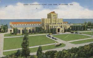 A painted postcard illustrates a bird's eye view of the grand building of Milwaukee's 1939 Water Purification Plant with Lake Michigan behind. The building has two wings. The central part is elaborate and features an entrance. Vehicles traverse a u-shape driveway around the entrance. Extensive green lawns appear in front of the building. Green trees span the foreground. Blue sky is above. Text at the top center reads "Water Purification Plant, Lake Park, Milwaukee, Wis."