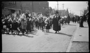 Grayscale wide shot of a crowd of children accompanied by women marching down Lincoln Avenue. Many children carry American stick flags. Some marchers carry signs urging the purchase of war bonds. Buildings and utility poles line the street side in the background.