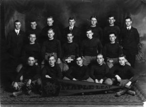 Grayscale group photo of the Maple Leaf amateur football team posing in a studio. They pose in three rows. Seven men stand in the back row. Five men sit in the second row. The person in the middle holds a ball that read "Milw. League Champions 1914." Five men in the front row sit cross-legged on the carpeted floor with a large "Maple Leaf" pennant flag.