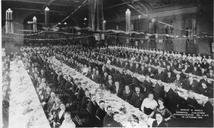 Grayscale elevated shot showing dozens of people sitting in rows of long dining tables in a large hall. They pose in formal attire and make eye contact with the camera lens. Women in identical dresses sit in a row in the right background. A group of people sit on the stage in the left background. Strands of ribbons ornament the ceiling and ceiling lights. Small flags appear here and there in the background.