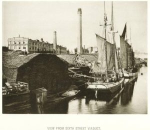Sepia-colored photograph of sailing boats lining the edge of a water body. The bank is on the left side. The Pfister and Vogel Tannery is seen in the left-to-center background. Text beneath the image reads "View From Sixth Street Viaduct."