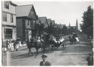 Grayscale long shot of two horse-drawn carriages parading on an empty street. Each four-wheeled carriage was pulled by two horses and carried a driver. Tall trees grow on the road verges. A group of people stands on the left side of the street, watching the parade. Behind them are multi-story buildings side by side as far as the eye can see.