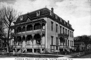 Long shot of facade and side of Morris Pratt Institute in grayscale tone. The two-and-a-half storybuilding has two entrances with stairs: one on the facade, one on the side. The facade features a balcony and a porch enclosed with ornamented balustrades and windows. The building's side has a decorated portico and regularly spaced rectangular windows. Dormer windows are set on the steep mansard roof. Text beneath the image reads "Morris Pratt Institute, Whitewater, WIS."