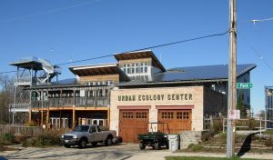 Exterior view of the Urban Ecology Center's main building facing slightly to the left. The building has a balcony and porch on the left and wooden garage doors on the right. Atop the doors is inscribed "Urban Ecology Center" in green. Two vehicles are parked in front of the garage. A pole with street signs appears in the right foreground. The blue sky is above.