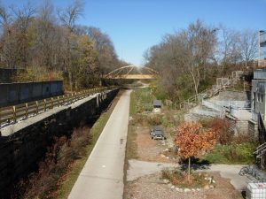 Elevated view of the Oak Leaf Trail stretching down next to the Urban Ecology Center. Another path stretches down on the left. Both tracks are separated by a fenced wall. A portion of the Center's building is visible on the right. East Park Place bridge appears in the distance above the trail. Trees can be seen here and there in the background.