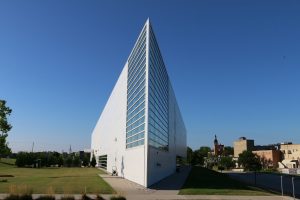 Long shot of the corner of the Museum of Wisconsin Art building. The image shows two sides of this triangular-shaped structure. Each side features a white-colored wall and a glass curtain wall that converge dramatically in the image's center. Green lawns appear outside the building on the left and right sides. Trees are seen on the left background. Other buildings are seen on the right back.