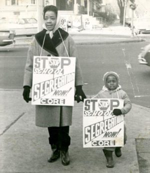 Grayscale full shot of a woman holding a child's hand in their warm clothes as they walk the sidewalk with protest signs hanging from their necks. The sign reads, "Stop School Segregation Now! CORE" The O in the word STOP is in the shape of a hexagon stop sign with the word STOP written in it.