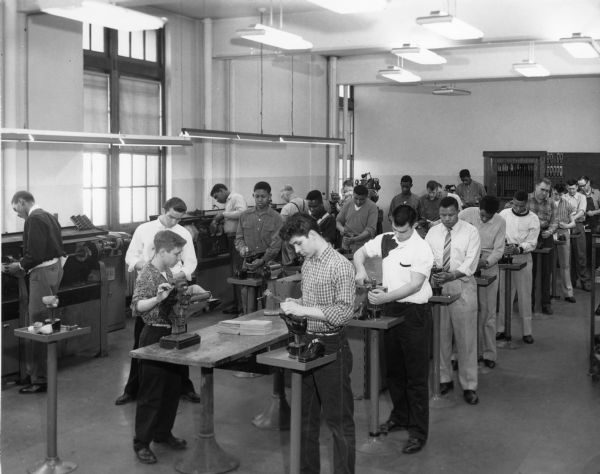 Rows of students in a shoe rebuilding class in a row of individual stations as they work on a shoe product. People on the left operate machines. The rows of students standing in the center and right are working with their hands. Students wear button-down shirts; one man wears a tie. The ceiling lights are on.