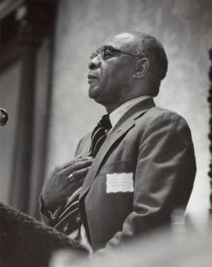 Grayscale portrait of Wesley Scott facing left in glasses, suit and tie. Scott stands with his right hand over his chest.