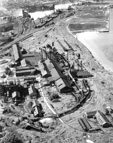 Aerial photo of the Bay View Rolling Mills showing a complex of buildings and rail tracks. It sits in a relatively low-density area. A body of water is located near the plant. Another building complex appears in the upper portion of the image