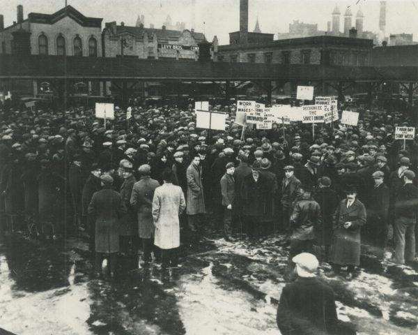 High-angle shot of a crowd of protestors standing close to each other in hats and warm coats. Some hold protest signs that demand workers' rights and recognition of the Soviet Union. The crowd is predominantly men.