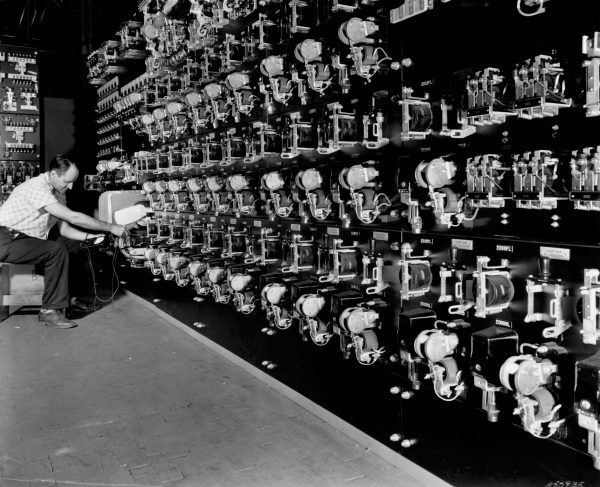 Grayscale wide shot of rows of electrical controls attached to a wall from left to right. A Cutler-Hammer worker tests one of the machines while sitting in a shirt and trousers.