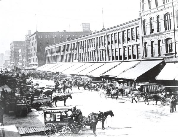 Grayscale wide shot of Broadway Street displaying long lines of horse-drawn vehicles parking next to the Commission Row's building complex.