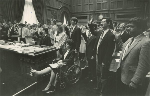 Long shot of a group of lawyers filling a courtroom, facing left while raising their right hands to take an oath. A long bench is on the farthest left in the foreground. On the center is a lawyer in a wheelchair with one leg up.