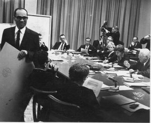 Photograph of Lloyd Barbee walking past a meeting table full of people sitting in formal attire with papers on the conference table in front of them. Near the top right of this grayscale image is a man working on a professional video camera that seems to be used to record the meeting. Barbee turns his back on these people while holding what looks like a large cardboard. He wears a dark suit and tie and round glasses.
