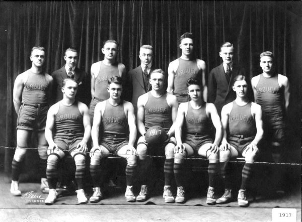 A grayscale group photo of Marquette Men's Basketball team in two rows. All five men sitting in the first row wearing uniforms that feature the word "Marquette." The one sitting in the middle holds a basketball that has the number "1917" painted on it. In the second row are four men in jerseys and three men in suits standing next to each other.