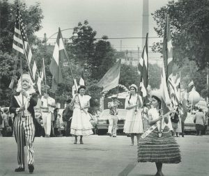 Sepia-colored full shot of a group of people in various costumes carrying different flags parading down the street during Mexican Fiesta. A man in the front left smiles while gazing up at the American flag he carries. Spectators are in the left background next to cars parked on the street side. More members of the crowd appear in the far right background. Trees grow on both sides.