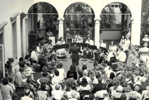 Sepia-colored high-angle shot of the back view of spectators watching the Milwaukee Chamber Orchestra performance in an outdoor space. Rows of audience members sit on chairs. Some stand and sit on the floor. The orchestra musicians play their instruments while seated in a semi-circle. The conductor is in the middle. Three arched openings and a balcony stand behind them.