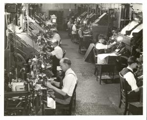 Sepia-colored photograph of two rows of employees working at intertype machines facing away from each other. Their chairs leave a narrow way in the middle where people can walk through.