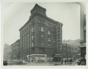 Sepia-colored long shot of Alhambra Theater on a street corner at the image's center. The building's corner on the ground floor features display windows of the Empress Hat Shops. Next to this is the Majestic Neckwear shop windows. The theater's entrance is on the right side. The marquee says, "Now Garden, Chester Morris, Frankie & Johnnie." Atop the marquee is a giant vertical sign that reads "Alhambra," attached to the multiple-story building's exterior walls. Adjacent buildings on the left and right sides are visible. Some people stand on the sidewalks. Text at the image's bottom reads "Alhambra Theater Building, W. Wisconsin Av."