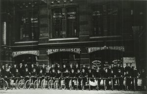 Grayscale long shot of bicycle-riding messengers posing in two rows in front of a building. All the young people in the front row pose in uniforms and bicycles. People in the back row stand in different clothes. Some are also in uniforms and holding bicycles. The building in the background has three bays. Three companies' signs are attached to each bay on the ground floor. The "Chicago Milwaukee Telegraph Co" sign is attached to the left bay. The "Henry Gollusch" sign is on the central bay. The "Western Union Telegraph" sign is on the right bay. Insurance company signs are visible on the second-floor's windows.