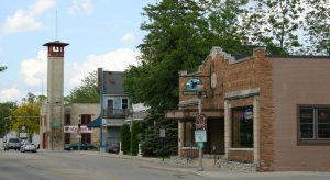 Long shot of the Main Street in Thiensville. The street stretches from left to right foreground. An intersection, cars, and traffic lights appear on the far left. Several buildings line the street side. Lush green bushes and trees are visible among the buildings in the image's center.