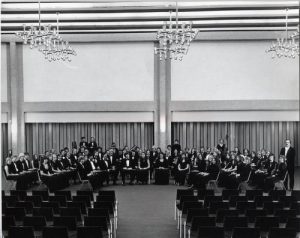 Grayscale group photo of the UWM Symphony Band sitting in three rows that form a semi-circle in the distance. Some people stand randomly behind the last row. Three chandeliers are on the ceiling. A tall wall is in the background. The rear view of rows of a seating area is in the foreground.
