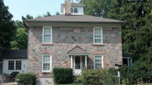 Facade of the Enoch Gardner and Mary Caroline Koch Needham House features fieldstone exterior walls. The two-story facade has four white-framed rectangular windows and a gabled entry porch in the middle. A cupola and a chimney are installed atop the roof. The building's one-story wings are visible on the left and right back. Tall green trees are in the far background. Landscaping plants and a lawn adorn the house's yard in the foreground.