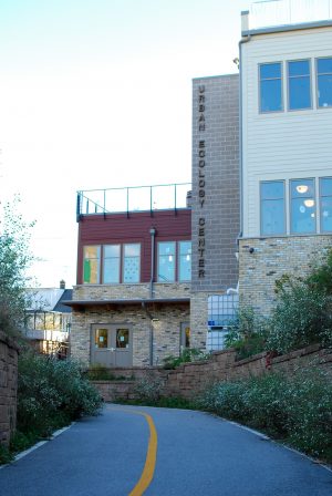 Side view of Urban Ecology Center facing left. The building's name signage is placed vertically on its exterior wall. In the foreground an asphalt road with a solid yellow line marks the center. A brown wall fence made of stone stretches along the right side of the road.
