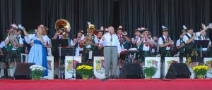 Wide shot of on-stage performance of musicians in traditional German garb. Most of them play an instrument. Two sing on a microphone while raising a glass. The conductor stands in the center.