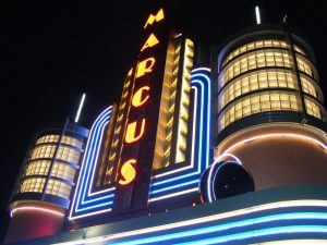 Low angle shot of the glowing Marcus Theatre marquee. A vertical sign that reads "Marcus" glows in yellow during the night time. Two small tower-like structures flank the sign. Both emanate lights.