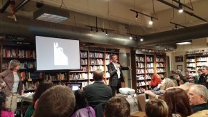 Interior of Boswell Book Company showing back view of audience members facing someone in a suit talking in the front. The man in the center background holds a mic with his right hand. On the farthest left is someone leaning on a table facing the audiences. Between the two people in the front is a projector screen. Behind them are a row of bookshelves from left to right.