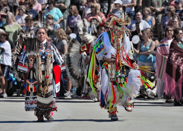 Long shot of two people in colorful cultural clothing dancing outside during a powwow in broad daylight. Beads, feathers, and ribbons embellish their clothes. The dancers' front bodies are visible. The face of person on the left is looking straight ahead. The other person lowers their head with one leg floating in a dance motion. Crowds stand in the background.