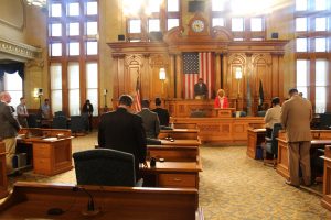 Back view of Common Council members standing in formal attire behind their desks in a chamber at City Hall. They stand toward the dais with a grand wooden wall panel that features an analog clock and an American flag in vertical position. Most people in the room bow their heads in prayer. Two people stand on the dais.