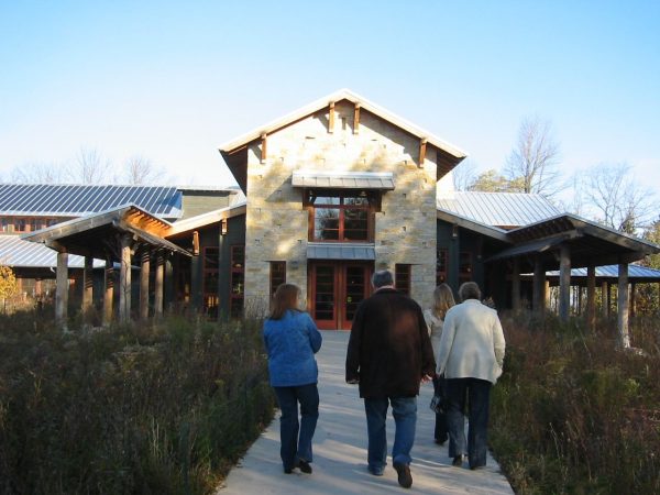 Exterior view of Schlitz Audubon Nature Center against the blue sky. Four people walk on a path toward the building's entrance. Green plants surround the left and right sides of the pathway.